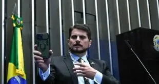 Homem de terno mostra a tela de um celular em plenário com painel escuro ao fundo