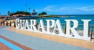 Letreiro branco com a palavra GUARAPARI em uma orla com mar e ilhas ao fundo