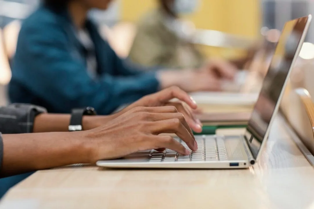 Mãos digitando em notebook sobre mesa, com outras pessoas estudando ao fundo