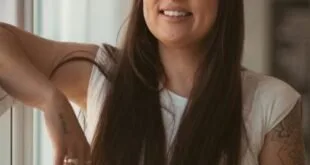Mulher de cabelos longos sorrindo apoiada na parede junto a uma janela