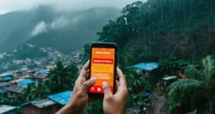 Mãos segurando celular com alerta da Defesa Civil na tela diante de encosta com casas sob chuva