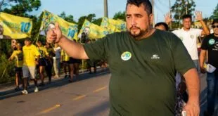 Homem de camiseta verde faz gesto de positivo em rua com bandeiras do Brasil ao fundo