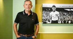 Homem de polo preta em pé ao lado de foto antiga de jogador de futebol na parede