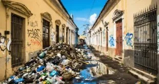 Montanha de lixo acumulado em rua estreita de casarões antigos com grafites e poça de água