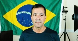 Homem de camiseta escura posa diante de bandeira do Brasil em estúdio com refletor e câmera