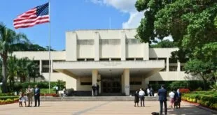 Fachada de embaixada dos Estados Unidos com bandeira hasteada e pessoas de terno no pátio