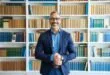Homem de barba grisalha e blazer azul sorri diante de estante repleta de livros