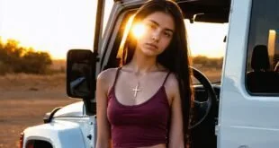 Jovem de blusa vinho encostada em picape branca ao pôr do sol em área de terra
