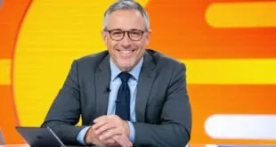 Apresentador de terno cinza sorri sentado à bancada de telejornal com fundo laranja