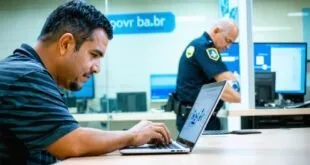 Homem digita em notebook em sala de atendimento com painel 'gov.br' e agente uniformizado ao fundo
