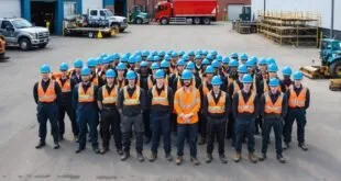 Grupo numeroso de trabalhadores de capacete azul e colete laranja perfilados em pátio com caminhões