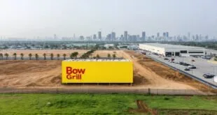 Vista aérea de terreno com painel amarelo, galpão industrial e horizonte de prédios ao fundo