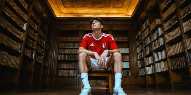 Jovem em uniforme esportivo vermelho sentado em biblioteca de madeira com prateleiras de livros
