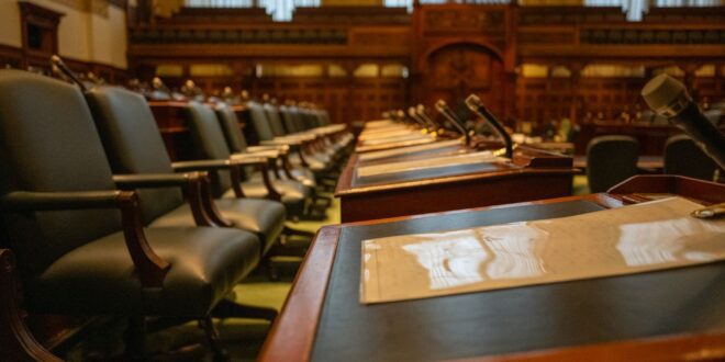 Cadeiras vazias e microfones nas bancadas de madeira de um plenário legislativo
