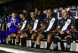 Jogadores sentados no banco de reservas de um estádio, usando uniformes preto e branco, conversando durante partida…