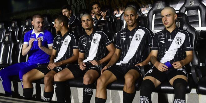 Jogadores sentados no banco de reservas de um estádio, usando uniformes preto e branco, conversando durante partida…