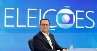 Homem de óculos e terno em estúdio de televisão, cenário azul, símbolo de pesquisas eleitorais