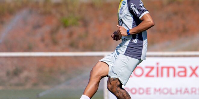 Jogador em uniforme azul e branco chutando bola em campo de futebol com arquibancada ao fundo