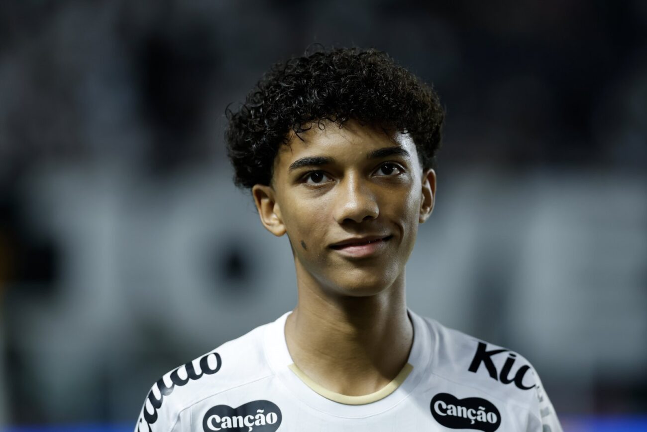 Jovem com cabelo preto encaracolado usando camiseta branca com inscrições, em close, com fundo desfocado