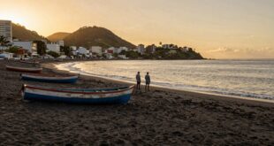 Duas pessoas em pé na praia ao entardecer, com barcos coloridos na areia e montanha ao fundo