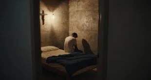Homem sentado em cama em cela com paredes de concreto, crucifixo na parede e luz focal