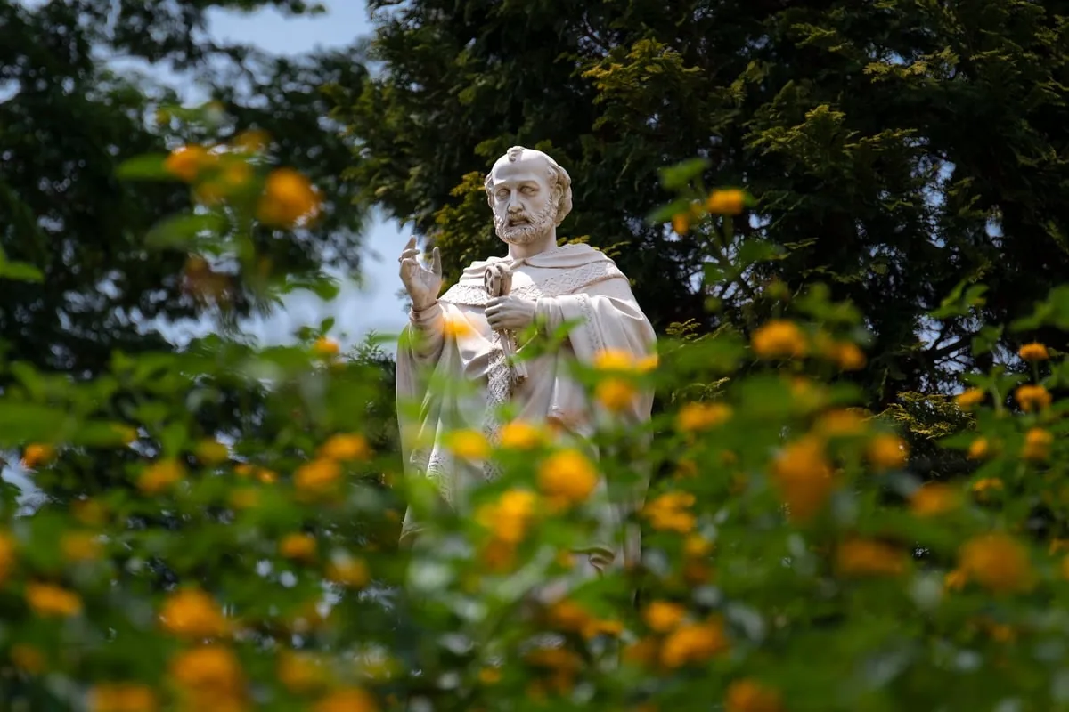 Estátua de frade em jardim cercada por pombos e um cachorro sentado ao lado