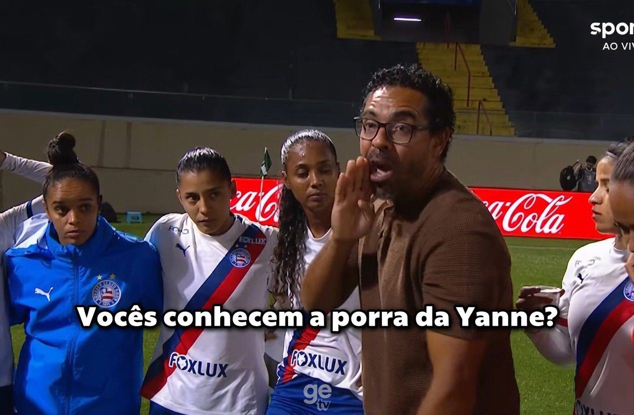 Homem de óculos e casaco marrom fala para grupo de mulheres em uniforme de futebol em campo com publicidade
