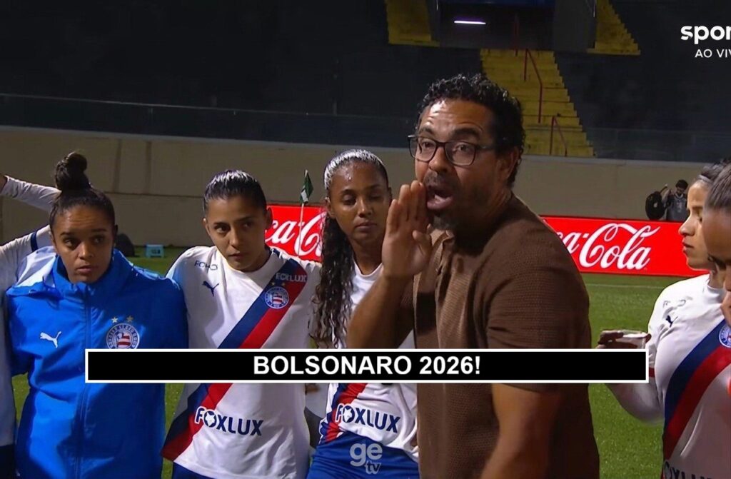 Homem de óculos e casaco marrom fala para grupo de mulheres em uniforme de futebol em campo com publicidade