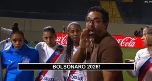 pênaltis: Homem de óculos e casaco marrom fala para grupo de mulheres em uniforme de futebol em campo com publicidade
