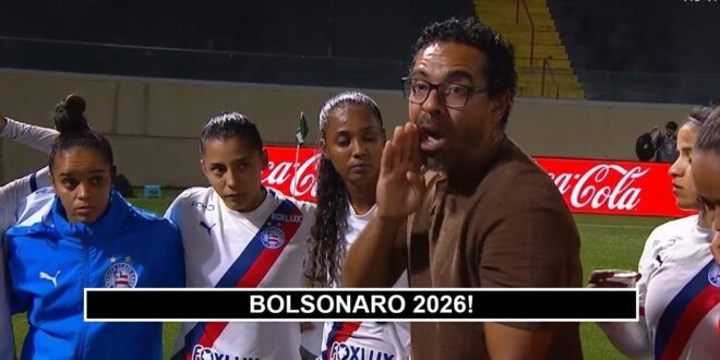 pênaltis: Homem de óculos e casaco marrom fala para grupo de mulheres em uniforme de futebol em campo com publicidade