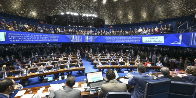 Parlamentares em sessão plenária em hemiciclo azul, com mesas, computadores, telão informativo e público nas galerias