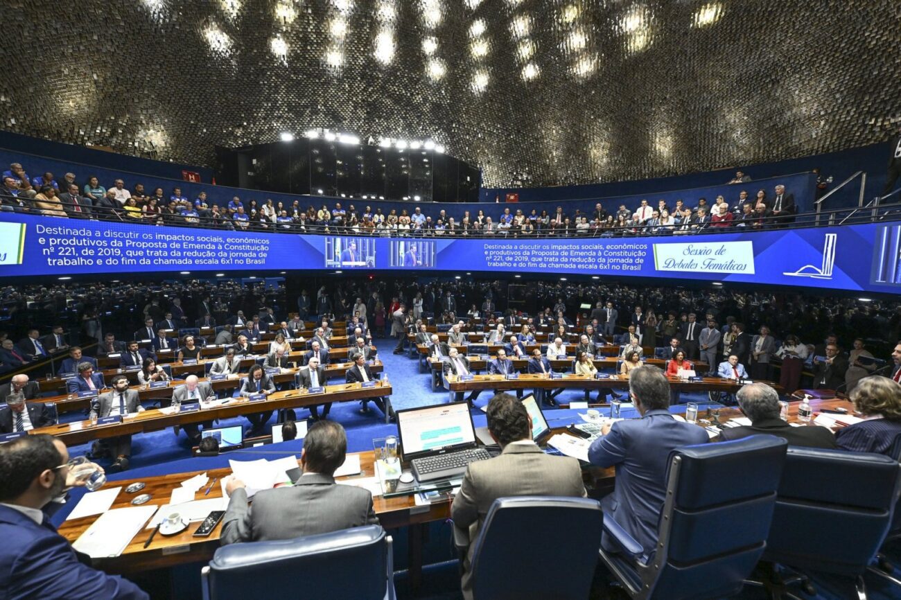 Parlamentares em sessão plenária em hemiciclo azul, com mesas, computadores, telão informativo e público nas galerias