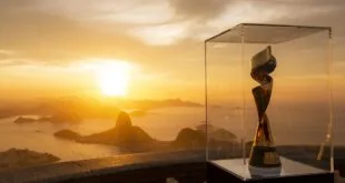 Troféu da Copa Feminina 2027 em vitrine com pôr do sol sobre baía e montanhas