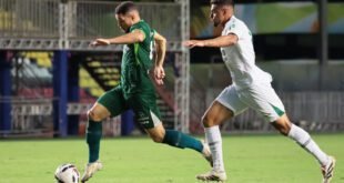 Jogador de verde conduz a bola perseguido por adversário de branco em campo iluminado