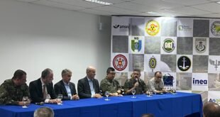 Autoridades civis e militares sentadas à mesa de coletiva diante de painel com logotipos