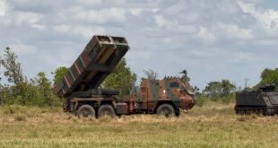 Lançador múltiplo de foguetes camuflado posicionado em campo aberto ao lado de blindado