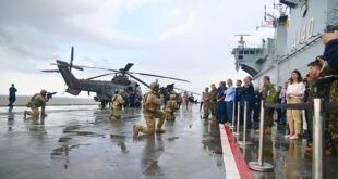 Militares armados ajoelhados no convés de navio de guerra observados por autoridades