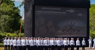 Militares em uniforme branco de gala perfilados diante de telão com o texto Dia do Aviador