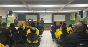 Pessoas de colete amarelo sentadas em sala assistindo a apresentação projetada