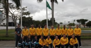 Equipe de atletas com agasalhos amarelos posa diante de mastro com a bandeira do Brasil