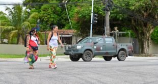 Duas mulheres atravessam rua diante de caminhonete militar com soldados na carroceria