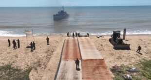 Navio militar ancorado próximo à praia com rampa de desembarque e militares na areia