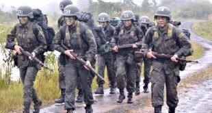 Militares com uniforme camuflado e mochilas marcham por estrada de terra sob neblina