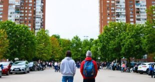 Duas pessoas de costas observam prédios residenciais altos enquanto moradores se reúnem na rua