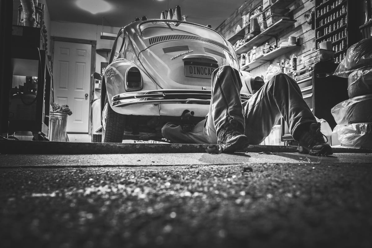 Mecânico trabalhando sob a lateral de um carro em oficina, em referência ao Dia do Mecânico