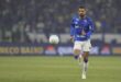 Jogador de futebol em uniforme azul e branco corre atrás da bola em campo durante partida