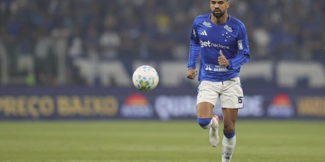 Jogador de futebol em uniforme azul e branco corre atrás da bola em campo durante partida