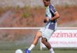 Jogador em uniforme azul e branco chutando bola em campo de futebol com arquibancada ao fundo
