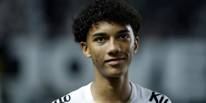 Jovem com cabelo preto encaracolado usando camiseta branca com inscrições, em close, com fundo desfocado